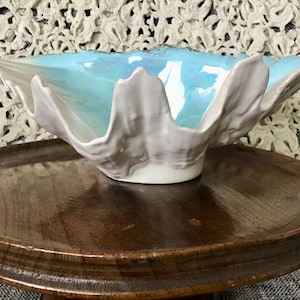May include: A decorative ceramic bowl with a scalloped edge, featuring a light blue iridescent interior and a white and gray exterior. The bowl is displayed on a round, wooden pedestal.