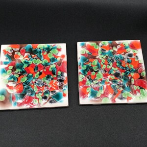 Può includere: Due sottobicchieri quadrati in ceramica con un design astratto colorato. I sottobicchieri presentano uno sfondo bianco con vortici e punti rossi, verdi e neri.