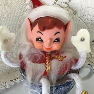 May include: A vintage Christmas elf doll with a smiling face, wearing a red and white outfit, white mittens, and a white fluffy collar. The elf is seated in a blue and white teacup.