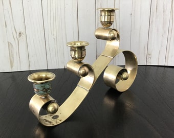 Könnte beinhalten: Ein goldfarbener Kerzenhalter aus Metall mit einem einzigartigen, fließenden Design. Er verfügt über drei Kerzenbecher und eine dekorative, wirbelnde Basis.