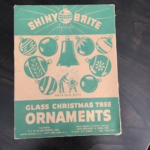 Könnte beinhalten: Vintage "Shiny Brite" Christbaumschmuck-Box. Die Box hat eine grüne und beige Farbgebung mit Illustrationen von Ornamenten und einer Glocke. Der Text enthält "American Made" und "Glass Christmas Tree Ornaments".