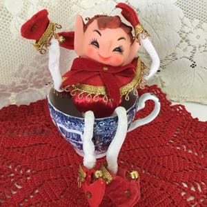 May include: A vintage Christmas elf doll sits in a blue and white teacup on a red crocheted doily. The elf wears a red velvet outfit with gold trim, a matching hat, and red shoes with gold bells. The elf's arms are white pipe cleaners.