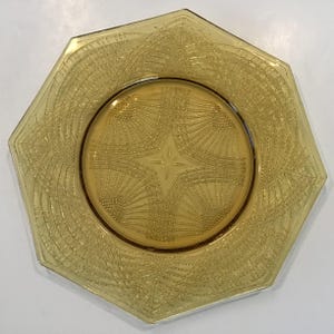 May include: A vintage amber-colored glass plate with an octagonal shape and a detailed etched design. The plate features a central starburst pattern and radiating lines, creating a textured surface. The plate's edges are slightly raised.