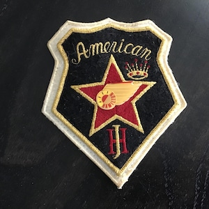 Op de afbeelding: Geborduurde patch met een zwarte achtergrond, een rode ster en gouden accenten. De patch heeft het woord "American" in gouden letters, een gouden vleugelontwerp en de naam "Jeff Hamilton".