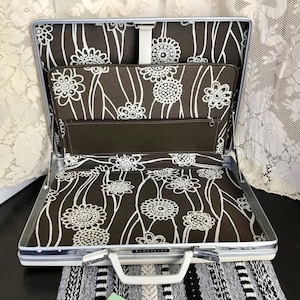 Puede incluir: Maletín Samsonite vintage abierto con marco plateado y asa blanca. El interior presenta un estampado floral marrón con flores blancas. El maletín tiene un bolsillo marrón y una correa blanca.