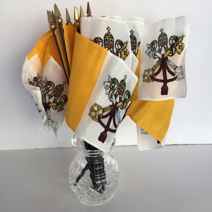 Può includere: Un mazzo di piccole bandiere della Città del Vaticano con colori giallo e bianco e lo stemma papale. Le bandiere sono su bastoncini neri e sono in un vaso di vetro trasparente.