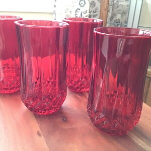 May include: A set of four tall, vibrant red drinking glasses. Each glass features a faceted, geometric diamond-cut pattern around the base that transitions into smooth, vertical panels toward the rim, creating a reflective surface.