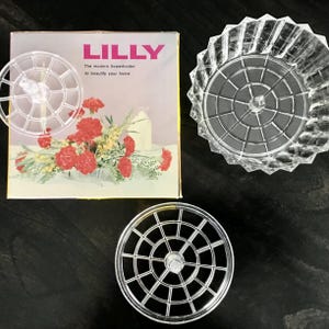 May include: Clear plastic flower holders and a decorative glass bowl are displayed on a dark surface. The box reads "LILLY" and "The modern flowerholder to beautify your home." The flower holders have a grid design.