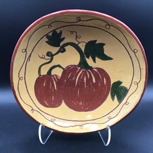 以下が含まれることがあります： 黄色い背景に、赤いカボチャ2つと緑のつるや葉が描かれた、手描きの丸い陶器の皿。縁は濃い赤色で、カボチャを囲むように装飾的なつるのデザインが施されています。