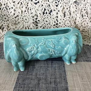 Pode incluir: Um vaso de cerâmica turquesa com formato retangular. O vaso apresenta detalhes em relevo de gnomos em cada extremidade e um desenho floral central. O vaso está sobre um tecido xadrez.