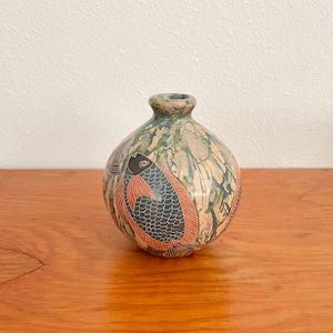 May include: A small, round ceramic vase with a narrow neck. The vase is decorated with a pattern of fish in black and orange against a mottled background of green, blue, and tan. The vase is sitting on a wooden surface.