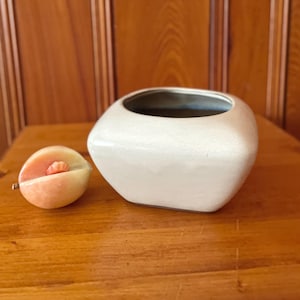 May include: A white ceramic square planter with a slightly rounded top and a smooth, matte finish. The planter is sitting on a wooden surface.