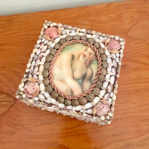 May include: A square-shaped trinket box decorated with seashells and a framed image of a woman. The box is made of wood and has a hinged lid.
