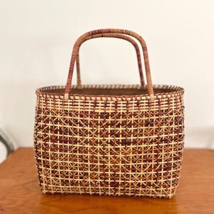 May include: A woven basket with a rectangular shape and two arched handles. The basket is made of natural-colored and reddish-brown woven material, creating a geometric pattern. The basket is sitting on a wooden surface.