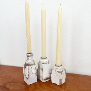 May include: Three off-white taper candles standing in individual square candle holders. Each holder features a distressed white painted finish over a wooden base, showing visible chips and wear. The candles are smooth, slender, and cylindrical.