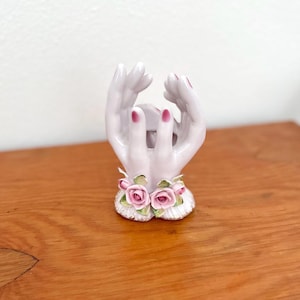 May include: A ceramic hand-shaped decorative piece. The hands are painted a pale lavender color with pink nail polish. A floral design of pink roses and green leaves adorns the base. The piece is set on a wooden surface.