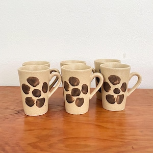 May include: A set of four beige ceramic mugs with a speckled texture, each featuring a dark brown painted flower motif consisting of six rounded petals. The mugs have a tapered cylindrical shape with a curved handle on the side of each piece.