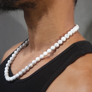 May include: A white beaded necklace with round beads. The necklace is worn by a person with a black tank top.