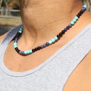 May include: A layered beaded necklace with black, blue, and turquoise beads. The necklace is worn by a person with a grey tank top.