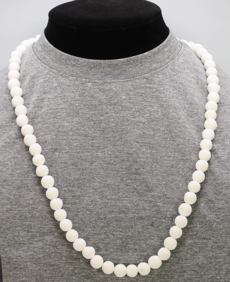 Puede incluir: Un collar largo de cuentas blancas hecho de cuentas redondas y lisas. El collar se muestra en una camiseta gris.