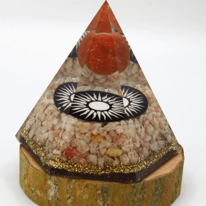 May include: A clear pyramid with a wooden base. The pyramid is filled with small pink and orange stones and has a black and white sun design on the side. The pyramid is about 6 inches tall.