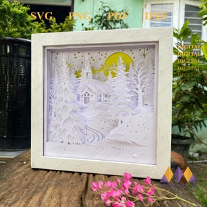 May include: A white papercut design of a winter scene with a church, trees, and a crescent moon. The design is framed in a white frame and is ready to be lit up.
