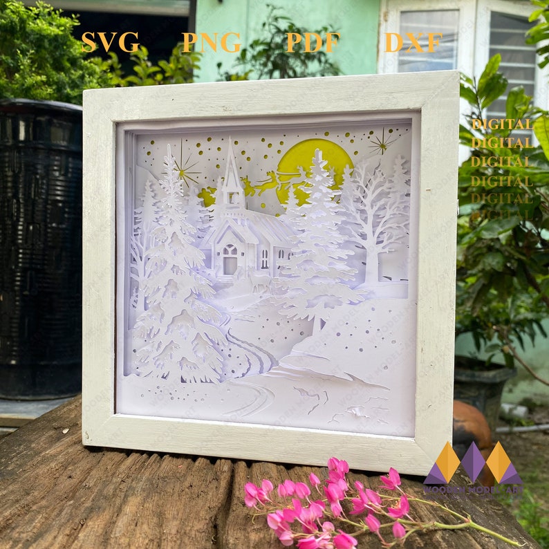 May include: A white papercut design of a winter scene with a church, trees, and a crescent moon. The design is framed in a white frame and is ready to be lit up.
