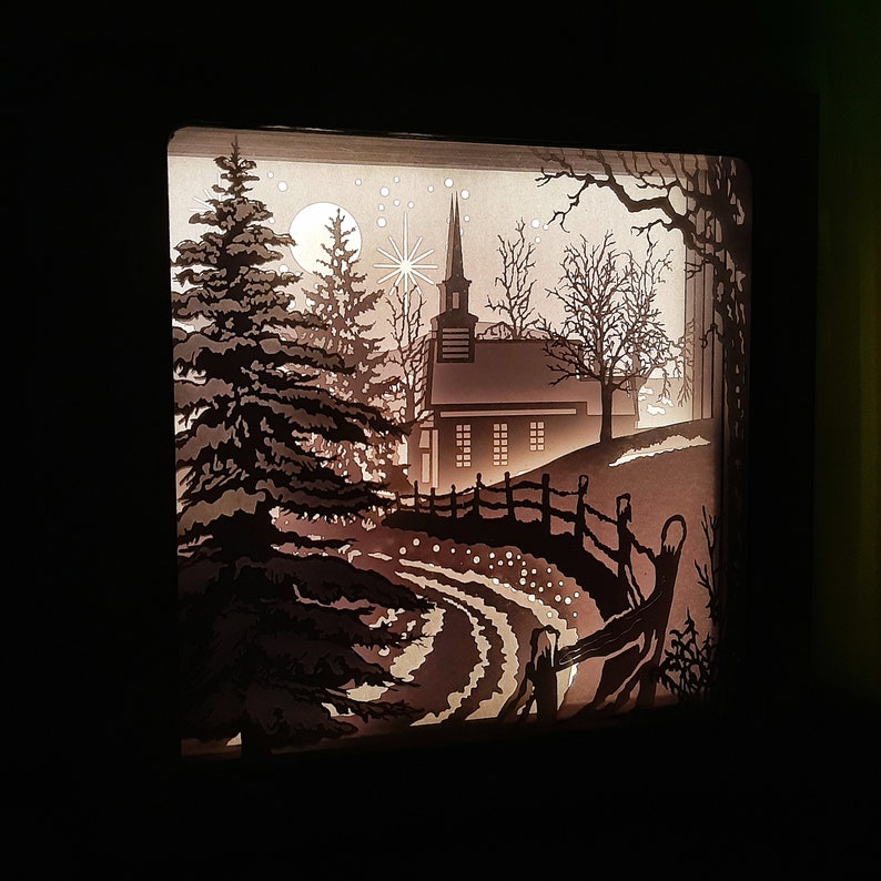 May include: A paper cut-out scene of a church with a steeple, surrounded by trees and a winding path. The scene is illuminated from behind, creating a silhouette effect.