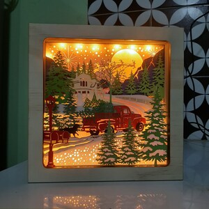 May include: A wooden light-up box with a winter scene featuring a red truck carrying a Christmas tree, a house, and snow-covered trees. The scene is illuminated with warm white lights.