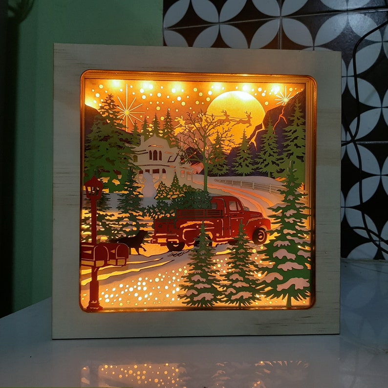 May include: A wooden light-up box with a winter scene featuring a red truck carrying a Christmas tree, a house, and snow-covered trees. The scene is illuminated with warm white lights.