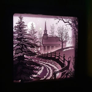 May include: A paper cut-out silhouette of a church with a steeple, trees, and a fence in a snowy landscape. The scene is illuminated with a soft, pink glow.