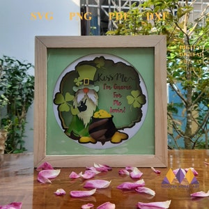May include: A wooden frame with a green background and a paper cut-out of a gnome holding a pot of gold. The gnome is wearing a green hat and has a white beard. The text "Kiss Me I'm Gnome For Me Lovin!" is written on the paper cut-out.
