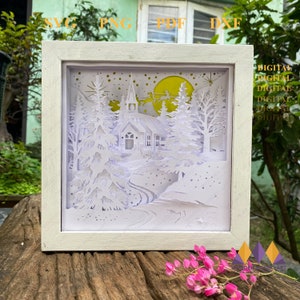 May include: A white papercut art piece in a white frame depicting a snowy winter scene with a church, trees, and a crescent moon. The scene is illuminated with a yellow glow. The words "DIGITAL DIGITAL DIGITAL DIGITAL DIGITAL" are visible on the right side of the image.
