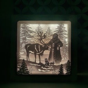 Puede incluir: Una caja de luz blanca con una silueta de Papá Noel, un reno, un gato y un conejo en una escena de bosque nevado.