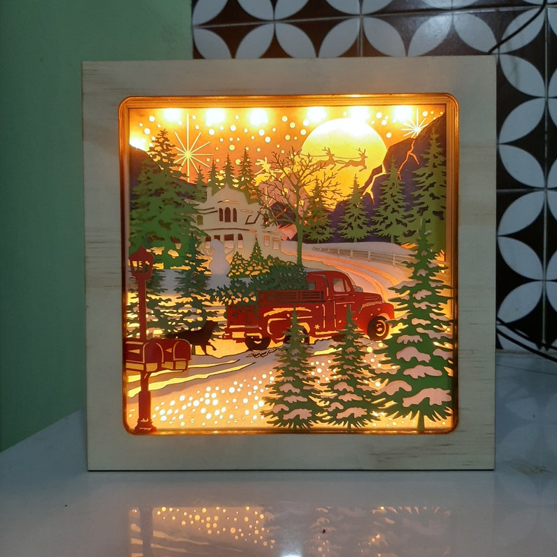 May include: A wooden light-up Christmas scene with a red truck carrying a Christmas tree, a house with a lit window, and a snowy forest landscape.