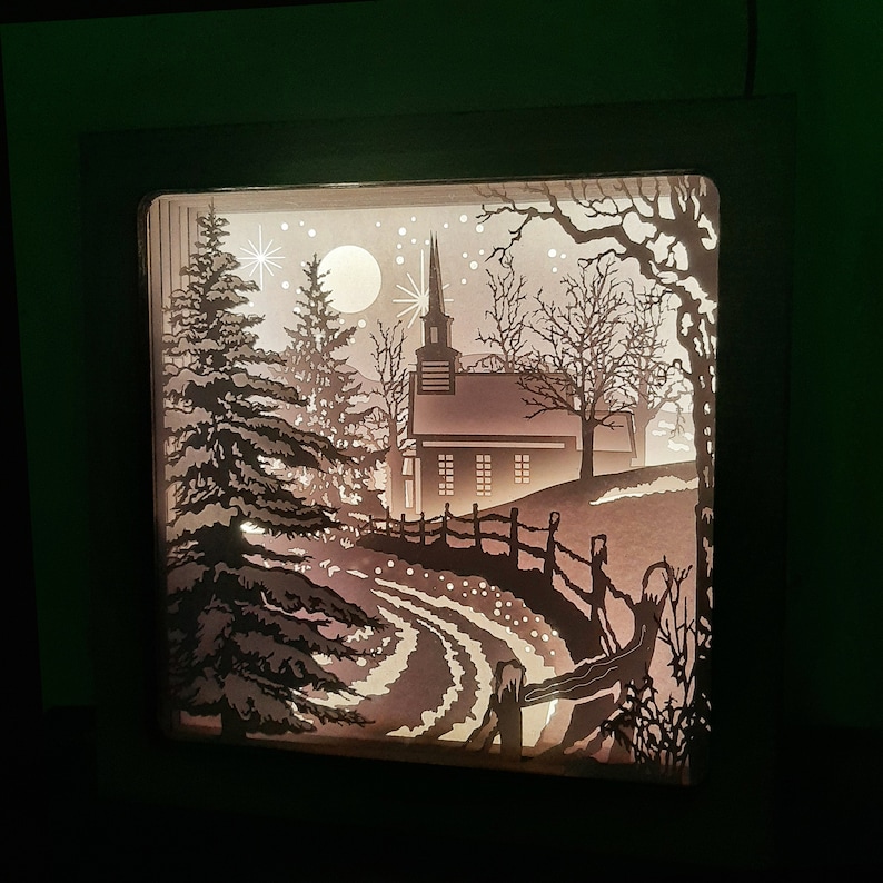 May include: A white silhouette of a church with a steeple and windows, surrounded by trees and a fence, is illuminated from behind. The scene is set against a dark background.