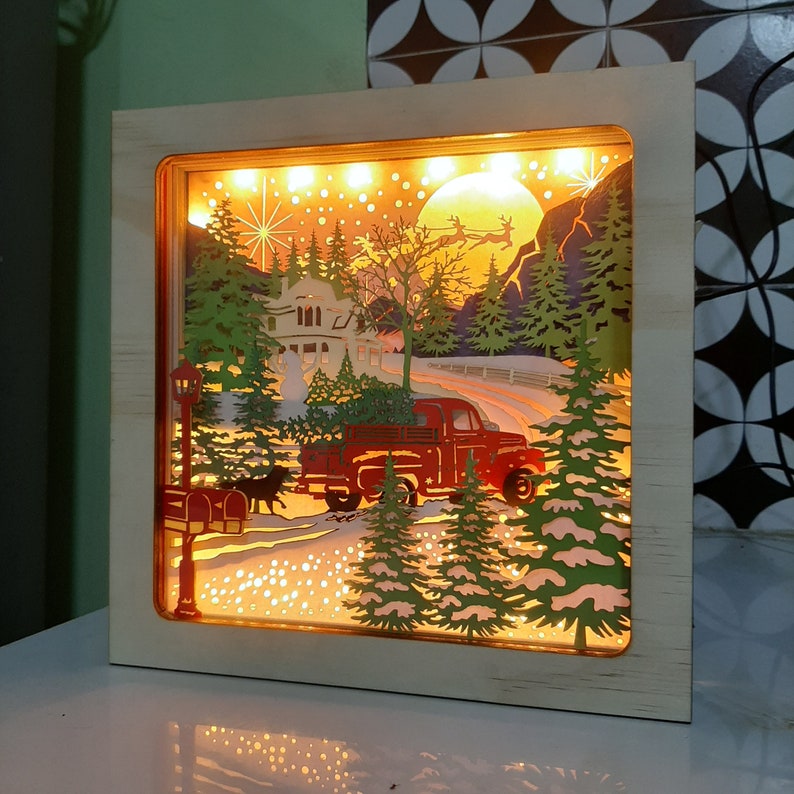May include: A wooden light-up box with a winter scene. The scene features a red truck with a Christmas tree in the back, a house with a lit window, and a snowy forest. The box is illuminated with warm white lights.