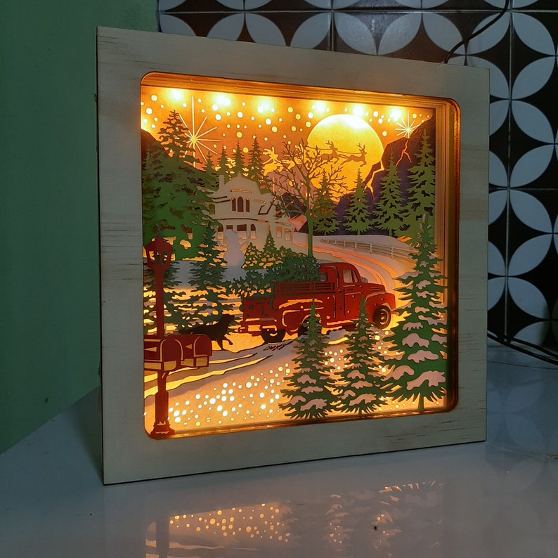 May include: A wooden light-up Christmas scene with a red truck carrying a Christmas tree, a house, and snow-covered trees. The scene is illuminated with warm white lights.