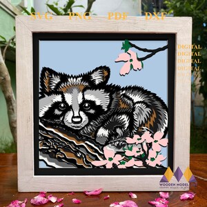 Könnte beinhalten: Eine digitale Schnittdatei mit einem Waschbären, der auf einem Baumstamm mit rosa Blüten sitzt. Das Design ist perfekt für die Erstellung von mehrschichtigen Papierarbeiten, wie z. B. Schattenboxen oder 3D-Kunst.