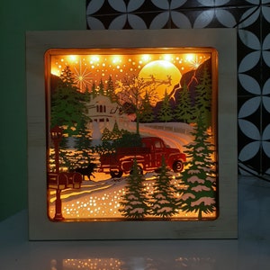 May include: A wooden shadow box with a winter scene featuring a red truck, a house, and trees. The scene is illuminated with white lights, creating a warm and festive glow.