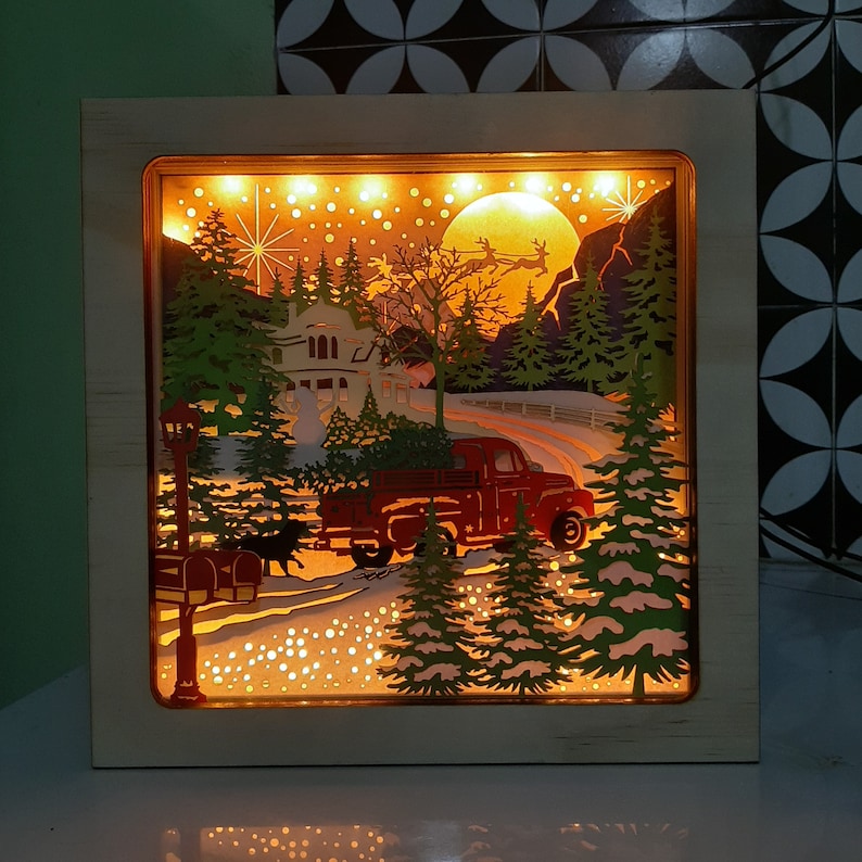 May include: A wooden shadow box with a winter scene featuring a red truck, a house, and trees. The scene is illuminated with white lights, creating a warm and festive glow.