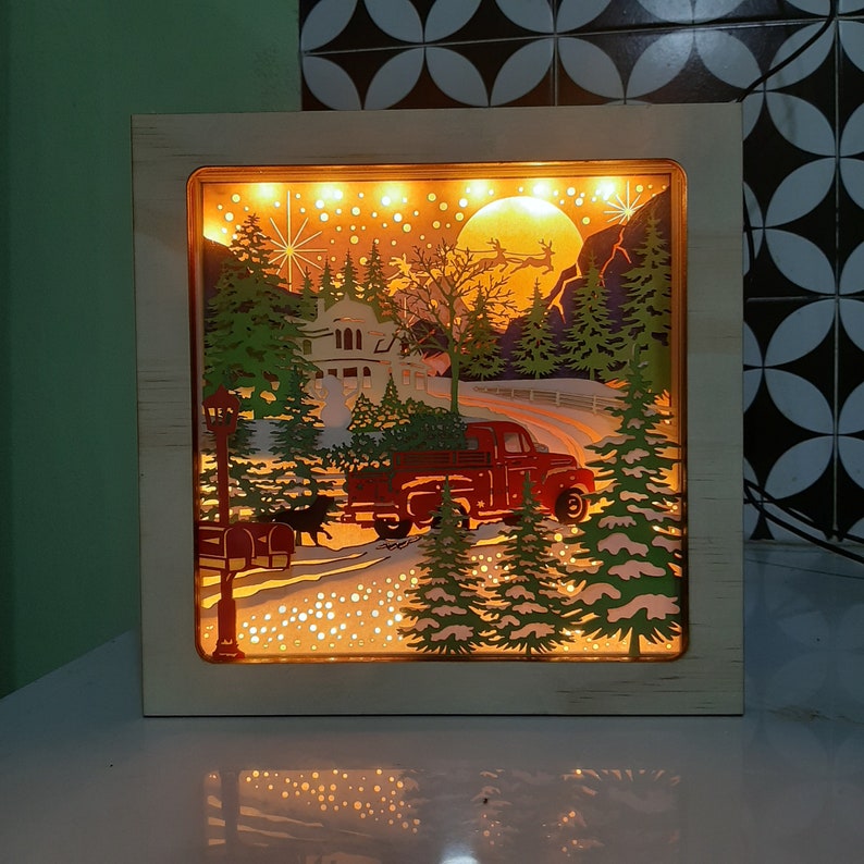 May include: A wooden light-up Christmas scene with a red truck, a house, and trees. The scene is illuminated with white lights and has a warm glow.