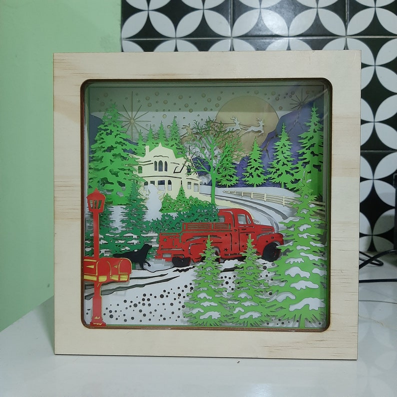 May include: A red truck with a Christmas tree in the back, driving through a snowy winter scene. The truck is surrounded by trees and a house with a white roof. The scene is illuminated by a bright moon and stars.