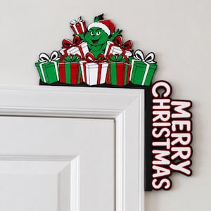 May include: A decorative door corner sign featuring a green character wearing a red Santa hat, surrounded by multiple wrapped gift boxes in red, white, and green. The vertical side of the sign displays the text MERRY CHRISTMAS in white letters.