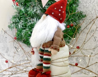 May include: A crocheted gnome doll with a red hat, white beard, brown sweater, and striped green, white, and red trousers. The gnome is sitting on a white surface.