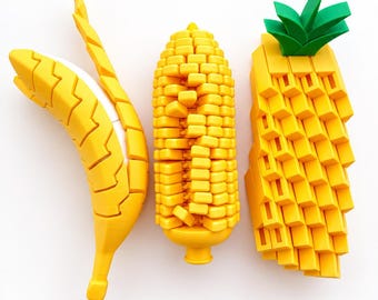 May include: Three articulated plastic figures shaped like fruit and vegetables: a yellow banana with a white core, a yellow corn cob with a textured surface, and a yellow pineapple with a green leafy top, all featuring a geometric block design.