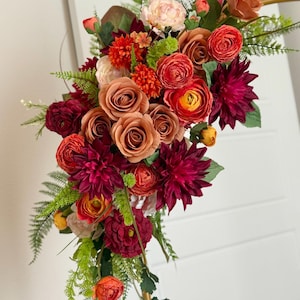May include: A vibrant floral arrangement featuring a mix of roses, dahlias, and other flowers in shades of burgundy, orange, and peach. Green fern-like foliage adds a natural touch. The bouquet is arranged on a gold metal stand.