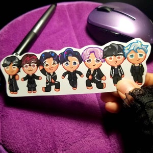 May include: A sticker featuring seven cartoon characters with various hair colors and styles, all wearing black outfits. The characters have a cookie-like design with smiling faces. The sticker is held against a purple background.