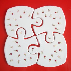Peut inclure: Quatre pièces de puzzle en céramique blanche avec un motif floral. Chaque pièce a une forme différente et présente de petites fleurs roses et rouges.
