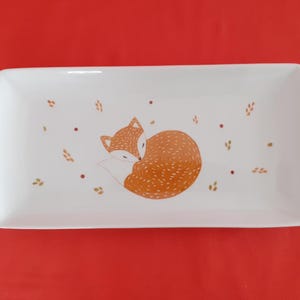 Peut inclure: Un plat de service rectangulaire en céramique blanche avec un motif peint à la main d'un renard orange enroulé avec des taches brunes. Le renard est entouré de petits motifs de feuilles brunes et dorées.